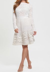 Kobieta ubrana w białą koronkową bluzkę z długim rękawem oraz spódnicę do kolan z haftem kwiatowym, zestawioną z beżowymi sandałkami na obcasie.