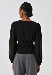 Blusa nera con maniche a palloncino e vita aderente, realizzata in un tessuto morbido. Abbinata a pantaloni grigi a vita alta, con dettagli arricciati sottili sul retro.