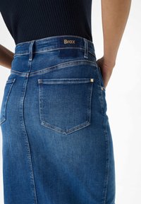 Blauwe denim rok met een getailleerd ontwerp, zichtbare achterzakken en gouden "Brax" branding op de tailleband. De textuur lijkt glad en duurzaam.