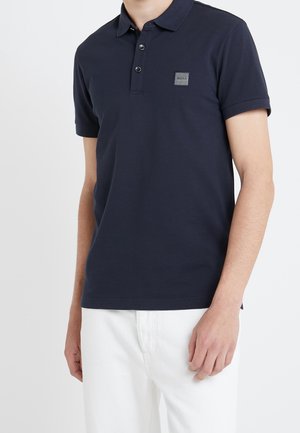 Homme portant un polo bleu marine à manches courtes avec trois boutons et un petit patch logo "BOSS" sur le côté gauche de la poitrine, associé à un pantalon blanc.