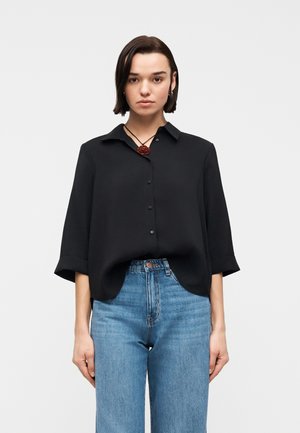 Kobieta z krótkimi ciemnymi włosami, ubrana w czarną koszulę na guziki z rękawami do łokci, niebieskie dżinsy z wysokim stanem i naszyjnik z czerwonej róży.