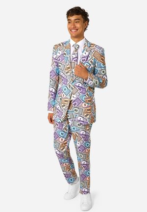 Jeune homme souriant, portant un costume coloré et une cravate recouverts de billets de Monopoly, debout avec une main près de sa poitrine.