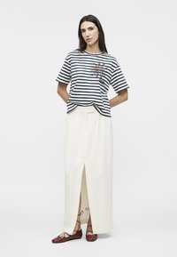 Femme portant un t-shirt rayé bleu marine avec un motif de soleil, une longue jupe blanche fendue à l'avant, et des ballerines brunes à lacets, debout devant un fond uni.