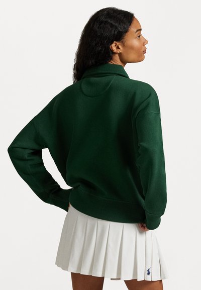 Green sweatshirt with a relaxed fit and high collar, featuring a soft texture and ribbed cuffs, paired with a white pleated skirt.