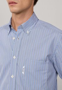 Hombre que lleva una camisa de botones a rayas azul y blanca con un bolsillo frontal y cuello abotonado.