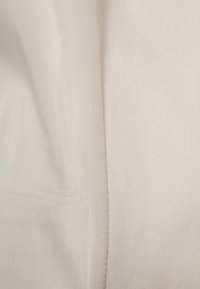 Světle béžová látka s hladkým texturem vykazuje jemné variace v lesku a má šev probíhající vertikálně po jedné straně.