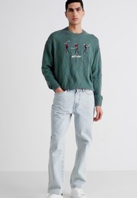 Groen gebreid sweater met een ruitpatroon, met geborduurde golfers en tekst. Gecombineerd met lichtblauwe spijkerbroek en witte sneakers.