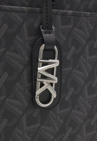 Sac à main noir en matériau texturé avec un motif répétitif, agrémenté d'un logo argenté et d'une bandoulière en cuir noir.