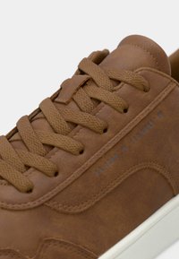 Brown leather sneaker with textured surface, flat laces, and a white rubber sole. Includes a discreet printed design on the side.