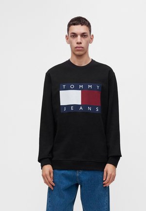 Νέος άνδρας που φοράει μαύρο φούτερ Tommy Jeans με λογότυπο και μπλε τζιν, στέκεται μπροστά σε απλό λευκό φόντο.