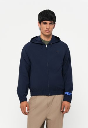 Navy blue zip-up jacket with a hood, elastic cuffs, and hem. Features a small logo on the chest and a blue accent label on the sleeve.