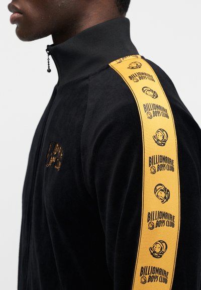 Μαύρο μπουφάν με φερμουάρ και ψηλό γιακά, διαθέτει κίτρινη πλευρική ρίγα με επαναλαμβανόμενο μοτίβο λογότυπων. Μαλακή υφή, εφαρμοστή σχεδίαση.