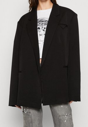 Persoon die een oversized zwarte blazer draagt over een wit grafisch crop top en grijze cargos met verfspatten, handen deels zichtbaar aan de zijkanten.