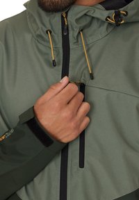 Groene waterdichte jas met een capuchon, voorzien van zwarte ritsen, verstelbare trekkoord en klittenbandsluitingen. Glad, mat stoffen textuur.