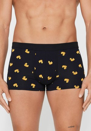 Torse masculin portant un boxer noir orné de petits canards jaunes flottants, debout les mains détendues le long du corps.