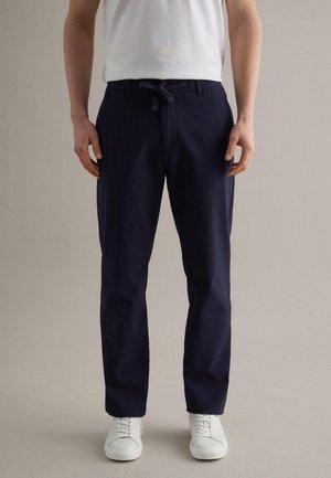 Uomo che indossa pantaloni blu navy con coulisse e trama, maglietta bianca e scarpe da ginnastica bianche, in piedi davanti a uno sfondo beige uniforme.