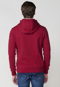 Felpa rossa con cappuccio e maniche lunghe, realizzata in un materiale morbido; presenta una tasca a marsupio, polsini e orlo a coste. Indossata con jeans blu.