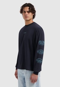 Langarm T-shirt in marineblauw katoen, met turquoise grafische tekst op de mouwen en borst. Losse pasvorm met geribbelde manchetten.