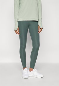 Leggings verdi realizzati in tessuto elasticizzato, con un design aderente e una texture liscia. Abbinati a una maglietta a maniche lunghe verde chiaro e sneakers bianche.