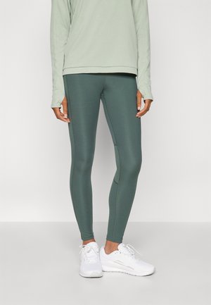 Leggings verdes hechos de tela elástica, con un diseño ajustado y una textura suave. Combinados con una blusa de manga larga verde claro y zapatillas blancas.