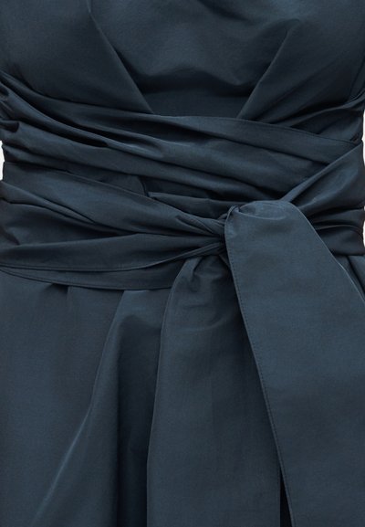 Σκούρο ναυτικό ύφασμα τυλιγμένο και δεμένο με φαρδιά κόμπο στη μέση, δημιουργώντας πτυχώσεις και ένα draped εφέ στα ρούχα.