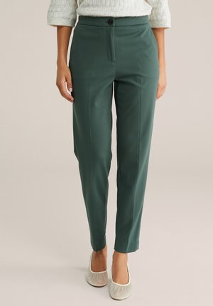 Femme portant un pantalon vert sur mesure, un pull en maille crème et des ballerines blanches, debout devant un fond uni.