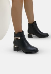 Botines negros de cuero sintético con un acento dorado en el tacón. Presentan un cierre lateral y un diseño de punta redonda.