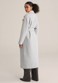 Palton lung gri din lână, cu talie cu curea, rever lat și mâneci lungi. Material texturat și croială până la genunchi. Poartă peste pantaloni închiși la culoare cu model.