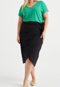 Grön texturerad v-ringad topp i kombination med en svart omslagskjol med sidoknyt. Bärs med ljusa vävda slip-on skor.