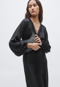 Čierne saténové šaty s dlhými nafúknutými rukávmi a čipkovaným výstrihom; modelka drží malú čiernu kabelku s korálkami a zakriveným tvarom.