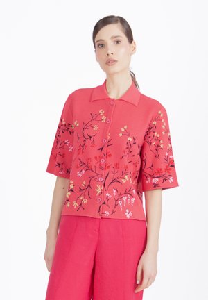 Mujer con camisa rosa brillante con bordado floral y pantalones de talle alto a juego, posando frente a un fondo blanco.