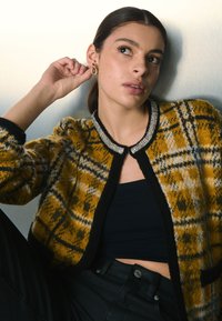 Cárdigan de cuadros amarillos y negros con ribete negro y escote con cuentas. La modelo lleva una camiseta negra y pendientes de aro. Fondo plateado.