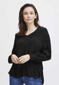 Melna blūze no mīksta auduma ar V veida izgriezumu, gariem pūkainiem piedurknēm un sānu šķirbm. Kombinēta ar ziliem džinsiem.