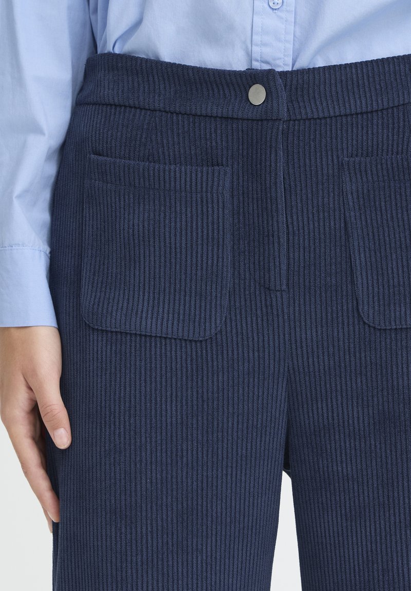 Gros plan sur un pantalon côtelé marine avec poches plaquées à l'avant et fermeture à boutons-pression, porté avec une chemise bleu clair à manches longues.