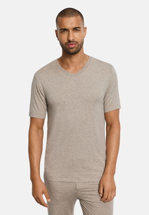 Mann mit kurzem Haar und Bart, der ein beiges, kurzärmeliges V-Ausschnitt-Hemd und passende Hose trägt, vor einem einfarbigen Hintergrund stehend.