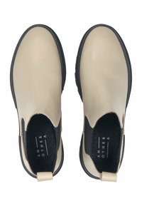 Béžové chelsea boty s lesklým povrchem, černými elastickými bočními panely a texturovanou černou stélkou. Obsahují poutko na opatku.