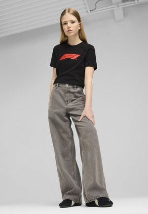 Čierne bavlnené tričko s červeným logom, kombinované s vysokými šedými nohavicami so širokými nohavicami. Model nosí čierne slip-on topánky. Jednoduché pozadie.