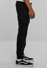 Pantalon cargo noir avec plusieurs poches, coupe fuselée, et poignets élastiques, assorti à des chaussures Vans noires avec des accents blancs.