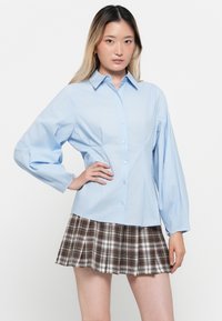 Camicia azzurro chiaro con maniche lunghe a sbuffo; abbinata a una mini gonna scozzese con tonalità marroni, bianche e grigie.
