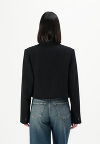 Svart kort blazer med långa ärmar, skräddarsydd passform och subtila axelpadder. Buren över ljusblå denimjeans med bakfickor.