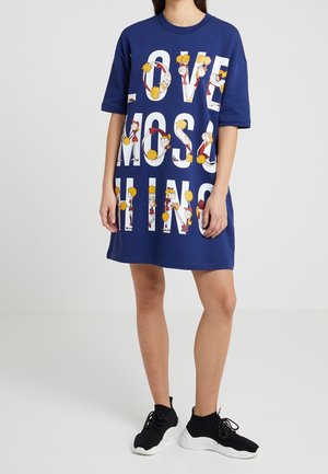 Marineblaues übergroßes Kleid mit "LOVE MOSCHINO"-Schriftzug, gefüllt mit Cartoon-Cheerleadern, die gelbe Pompons halten, getragen mit schwarzen und weißen Turnschuhen.