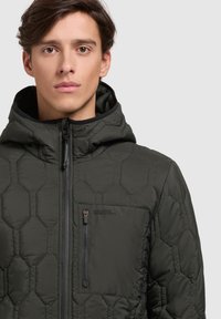 Dark green puffer jacket with hexagonal quilted pattern, featuring a zippered front pocket, hood, and black trim around the collar.