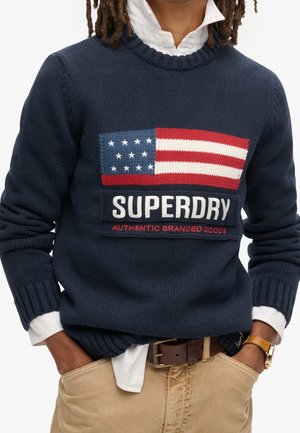Person iført en navyblå sweater med et amerikansk flagdesign og teksten "SUPERDRY AUTHENTIC BRANDED GOODS", kombineret med beige bukser og et ur.
