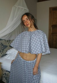 Γυναίκα με μπλε καρό crop top και αντίστοιχη φούστα στέκεται κοντά σε κρεβάτι με λευκό δίχτυ ουρανό σε απαλά φωτισμένο δωμάτιο.
