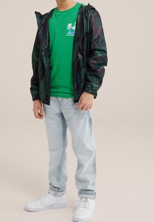 Black and green camo hooded jacket, green graphic t-shirt with palm trees, light blue jeans, and white sneakers. Smooth fabric, casual fit.