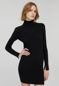 Abito nero a costine con collo alto, maniche lunghe e fessure laterali. Il tessuto ha una consistenza morbida e una silhouette aderente.