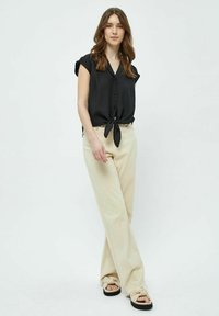 Schwarze kurzärmelige Bluse mit Knopfschluss und gebundenem Vorderteil, kombiniert mit hochgeschnittenen, cremfärbigen weiten Hosen und beigen, geknoteten Sandalen.