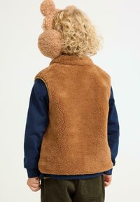 Braun flauschige Fleece-Weste mit hohem Kragen, kombiniert mit einem marineblauen Langarmshirt. Hat lockige Haare und rosa warme Ohrenschützer.