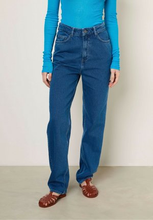 Person wearing high-waisted blue jeans, turquoise long-sleeve top, and brown closed-toe woven leather sandals standing on a neutral floor.