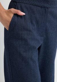 Hand met verzorgde nagels in de zak van een marineblauwe pinstripe broek, toont de stoftextuur en pasvorm.